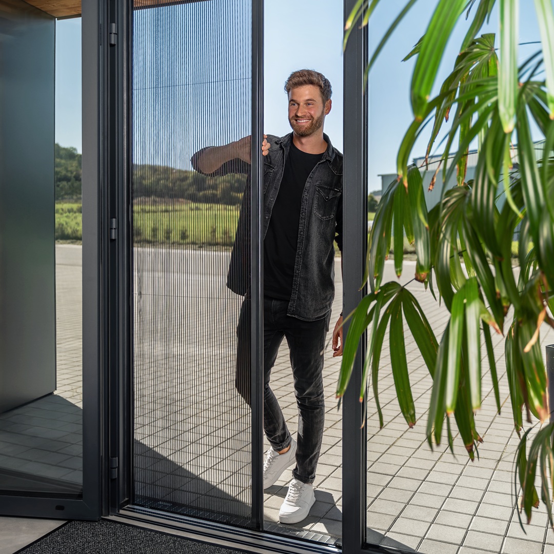 Ein Mann betritt ein Gebäude durch eine Tür. Er nutzt ein Fliegengitterplissee und lacht. Im Vordergrund ein moderner Baum mit langen Blättern.