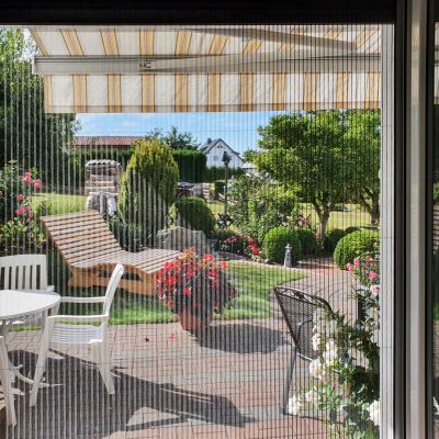 Fliegengitterplissee geschlossen an Terrassentür. Dahinter eine Markise und ein Garten.